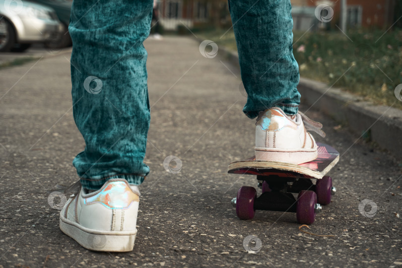 Скачать Маленькая девочка стоит на парковке в Асвальте и учится кататься на скейтборде. Девушка в джинсах, белой футболке и белой краске с желтым браслетом фотосток Ozero