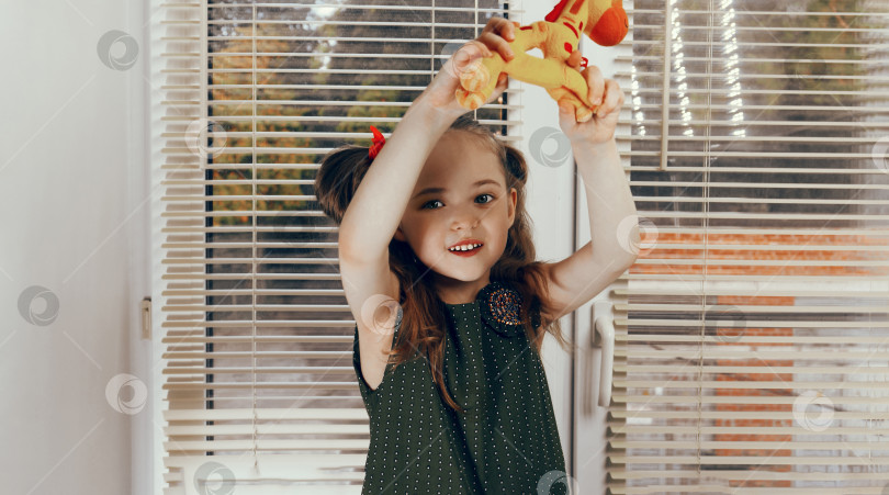 Скачать Маленькая девочка в темно-зеленом длинном платье ручной работы, с густыми хвостиками, держит игрушечного желтого жирафа. На заднем плане окно с жалюзи фотосток Ozero