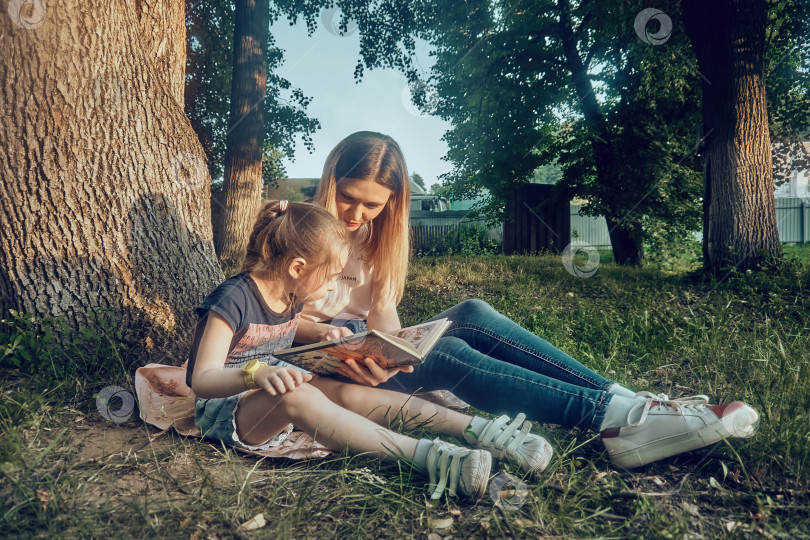 Скачать Мать и дочь читают книгу у дерева солнечным летним днем. Открытый парк фотосток Ozero