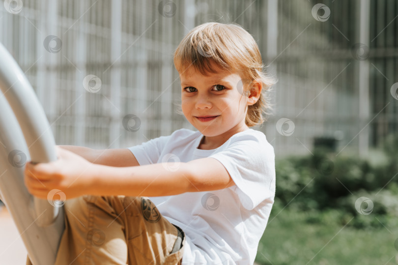Скачать ребенок на городской детской площадке качается на карусели. милый маленький счастливый улыбающийся откровенный пятилетний мальчик с длинными светлыми волосами в белой футболке. поколение z дети психическое здоровье образ жизни фотосток Ozero