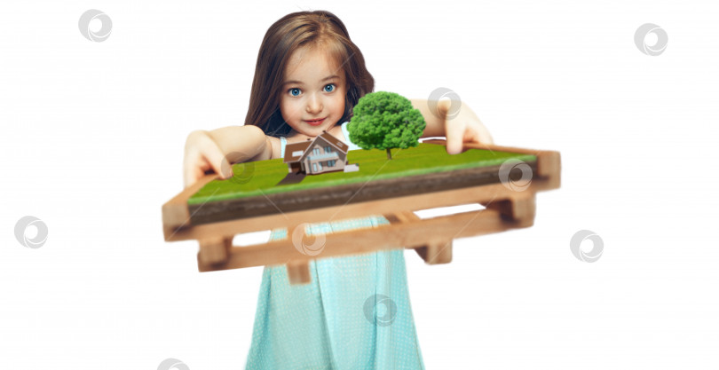 Скачать Маленькая девочка в голубом платье на белом изолированном фоне держит макет. Дом стоит на зеленой лужайке, а рядом растет большое фруктовое дерево. Выборочный мягкий фокус фотосток Ozero