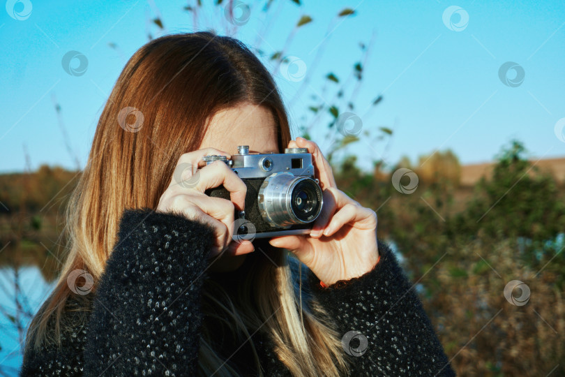 Скачать Девушка в черном пальто держит и фотографирует старым пленочным фотоаппаратом серебристо-черного цвета. Голубое небо на заднем плане фотосток Ozero