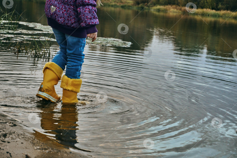 Скачать Ребенок в желтых резиновых сапогах, синих джинсах и фиолетовой куртке стоит в воде, не обращая внимания на погоду. В резиновых сапогах в воде фотосток Ozero