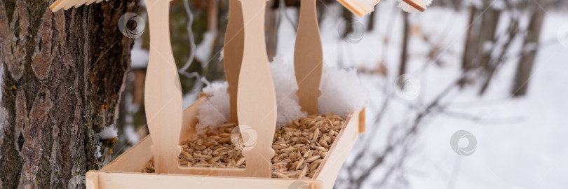 Скачать деревянная кормушка для диких лесных птиц с кормом, висящая на дереве, покрытом свежезамороженным снегом и снежинками в морозный зимний день в лесу или саду. уход за животными. снежный зимний сезон на природе фотосток Ozero