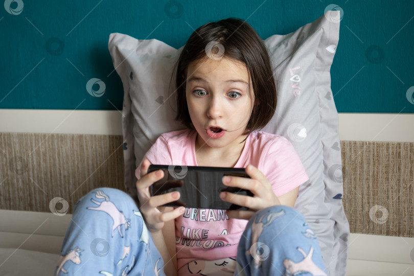 Скачать Ребенок сидит в постели и очень эмоционально смотрит в свой смартфон фотосток Ozero