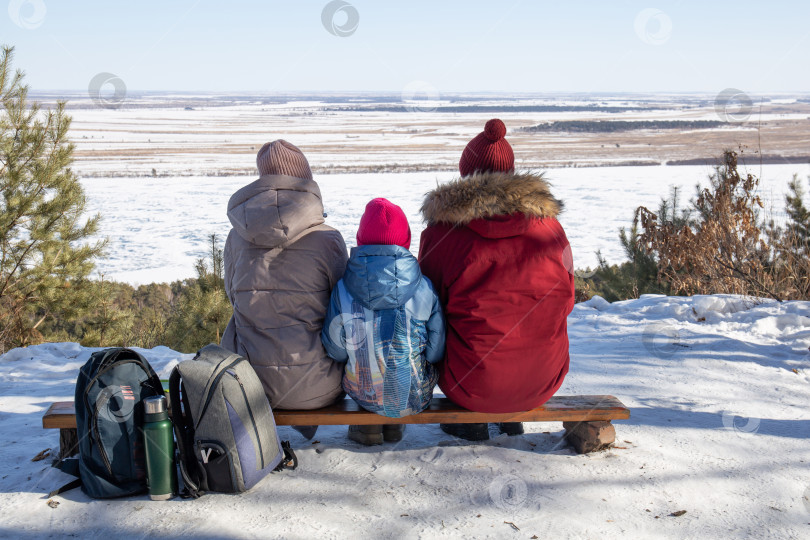 Скачать Семья в зимних пуховиках сидит лицом к камере на скамейке на вершине горы и смотрит вдаль. фотосток Ozero