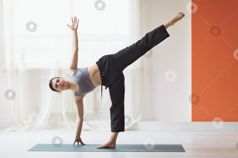 Скачать Молодая женщина, практикующая йогу, выполняет упражнение Ардха Чандрасана, позу полумесяца в студии у окна фотосток Ozero