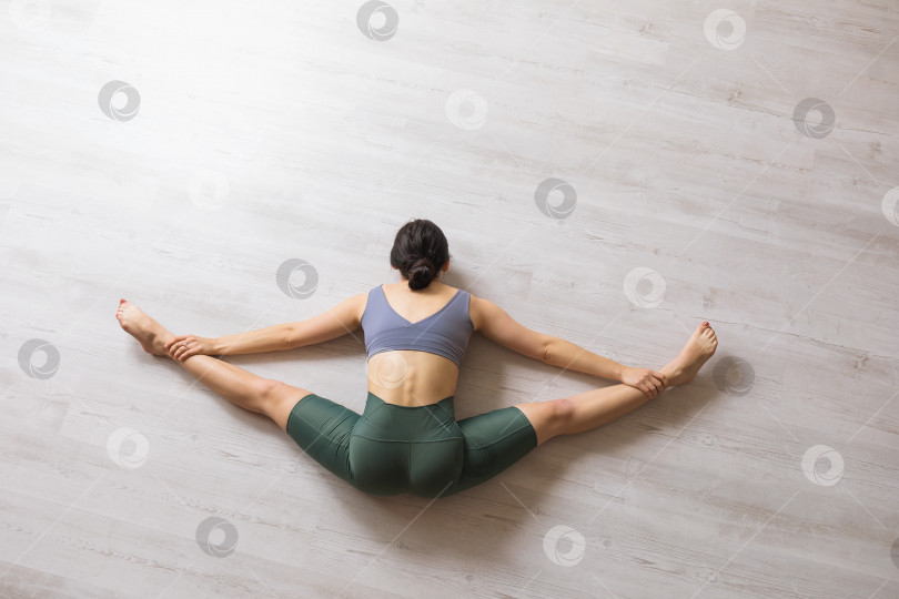 Скачать Женщина в спортивных леггинсах и короткой футболке, практикующая йогу, выполняет упражнение самоканасана, сидя на полу, поперечный шпагат с наклоном тела вперед фотосток Ozero