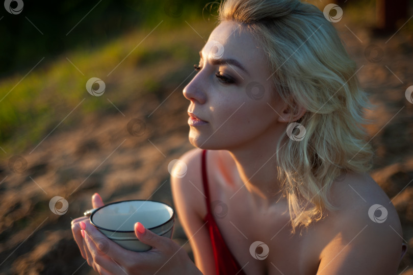 Скачать Красивая девушка, укрытая пледом вечером на закате, держит кружку чая двумя руками на фоне моря. фотосток Ozero