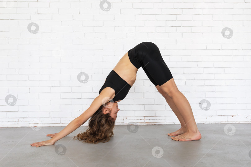 Скачать Молодая женщина, ведущая здоровый образ жизни и практикующая йогу, выполняет упражнение Адхо Мукха Шванасана, позу собаки лицом вниз, тренируется в черной спортивной одежде возле кирпичной стены фотосток Ozero