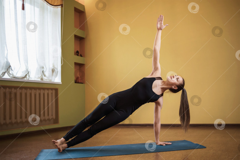 Скачать Женщина европейской внешности, практикующая йогу, выполняет упражнение васиштхасана, позу боковой планки, носит цельную спортивную одежду черного цвета, в помещении в полный рост фотосток Ozero