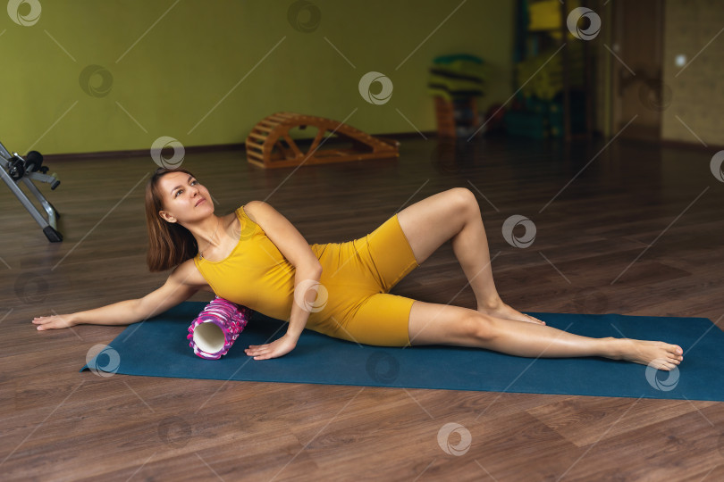 Скачать Женщина в спортивной одежде выполняет миофасциальный массаж боковых мышц спины и груди с помощью валика, лежа на коврике в комнате фотосток Ozero