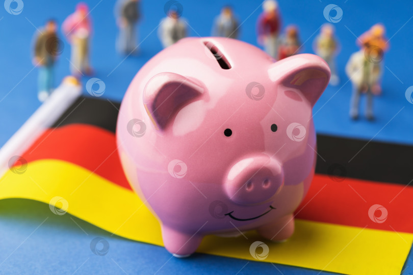 Скачать Копилка, флаг Германии и пластмассовые игрушечные человечки на цветном фоне, концепт на тему доходов населения Германии фотосток Ozero