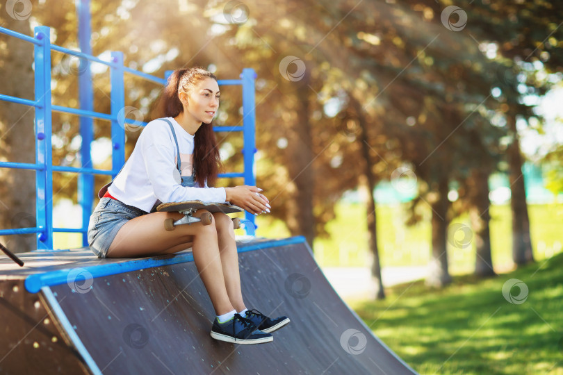 Скачать Молодая шатенка отдыхает со скейтбордом в руках перед катанием в парке фотосток Ozero
