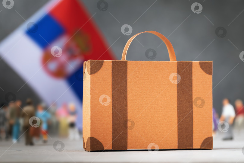 Скачать Картонный чемодан, пластмассовые игрушечные человечки и флаг на абстрактном фоне, концепция на тему переезда или миграции в Сербию фотосток Ozero