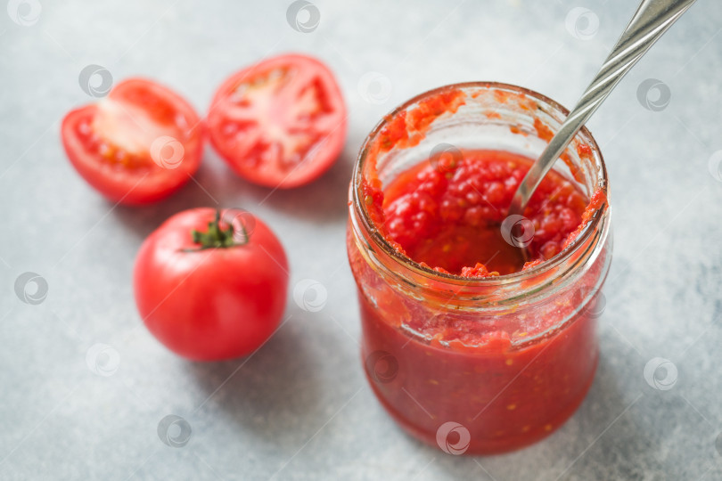 Скачать Томатная паста, консервированная в стеклянной банке, и свежие помидоры на бетонном фоне. Ферментация продуктов. фотосток Ozero