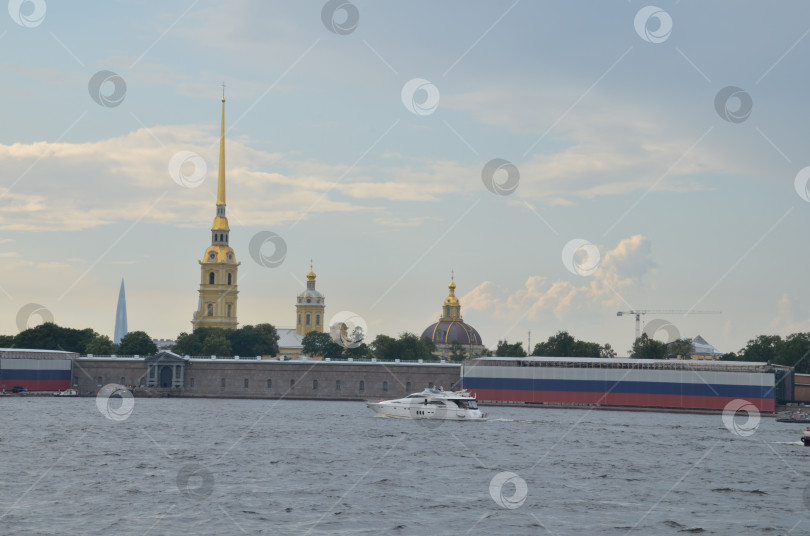 Скачать Санкт-Петербург, Россия – 07.27.2024: Петропавловская крепость на Заячьем острову в Санкт-Петербурге, крепостные стены украшены растяжкой в цветах флага Российской Федерации, мимо плывет белая яхта фотосток Ozero