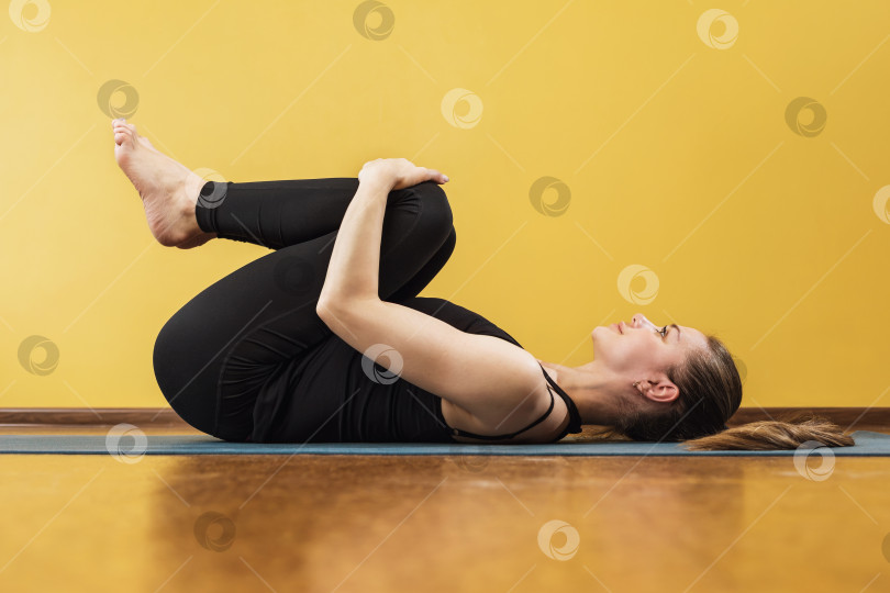 Скачать Спортивная привлекательная женщина, практикующая йогу, выполняет упражнение апанасана на коврике у стены, поза освобождения от ветра, в студии йоги фотосток Ozero