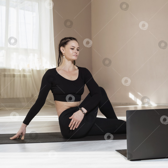 Скачать Женщина в черной спортивной одежде, сидящая на коврике и пристально смотрящая на экран ноутбука, концепция онлайн-тренировки фотосток Ozero