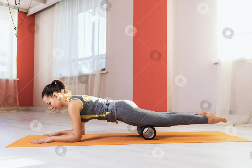 Скачать Женщина в спортивной одежде выполняет миофасциальный массаж мышц бедра с помощью валика на коврике в тренажерном зале фотосток Ozero