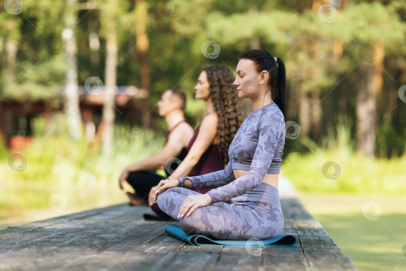 Скачать Три человека, практикующие здоровый образ жизни, занимаются медитацией, сидя на коврике в позе лотоса на деревянном мосту в парке летним солнечным утром фотосток Ozero