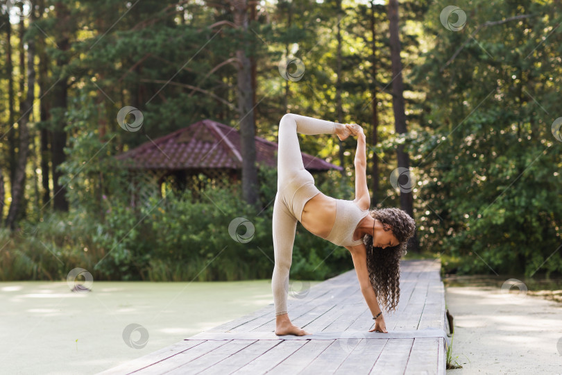 Скачать Женщина, практикующая йогу, выполняющая упражнение ардха чандрасана, поза полумесяца, стоит на деревянном мосту в парке солнечным летним утром, тренируется в спортивных леггинсах и коротком топе фотосток Ozero