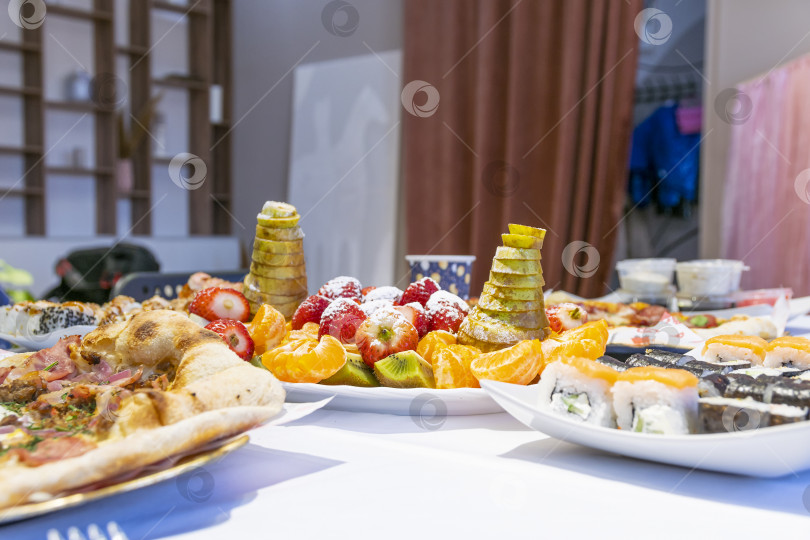 Скачать Банкетный стол для празднования детского дня рождения. На белом столе стоят тарелки с фруктовой нарезкой, суши и пиццей. фотосток Ozero