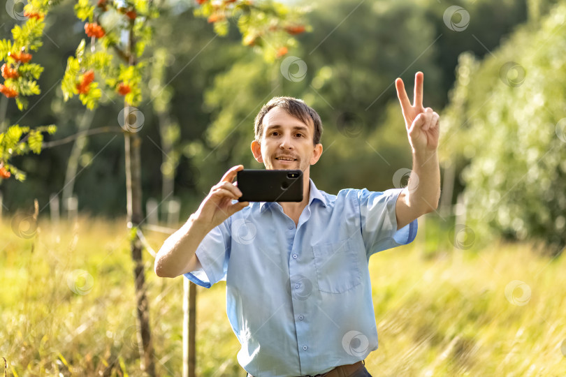 Скачать Молодой человек, уверенно улыбаясь, делает фото на смартфон в парке фотосток Ozero