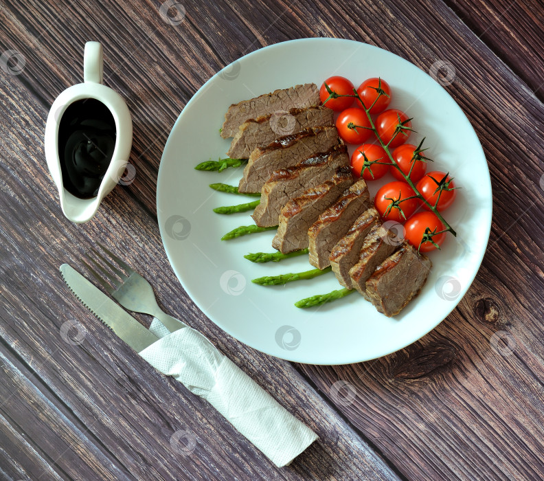 Скачать Приготовленный на гриле говяжий стейк, нарезанный кусочками, с бисквитом и помидорами черри выложите на деревянный стол рядом с соусом и столовыми приборами в салфетке. фотосток Ozero