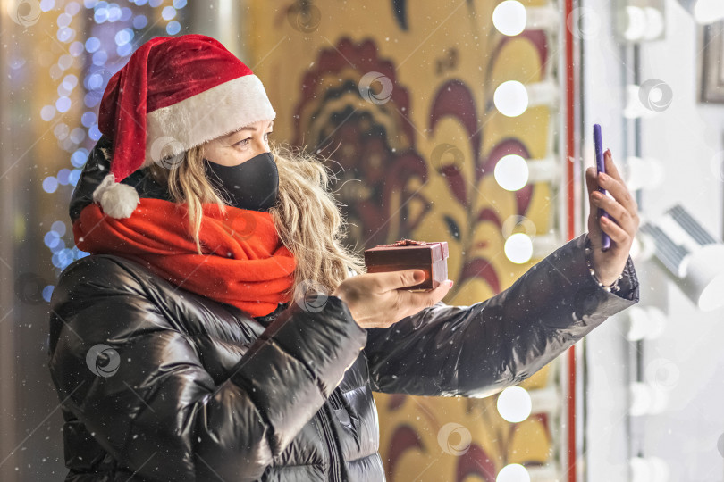 Скачать Блондинка средних лет в шляпе Санта-Клауса и защитной маске поздравляет друзей с Рождеством с помощью видео на смартфоне. Рождественские каникулы,шопинг фотосток Ozero
