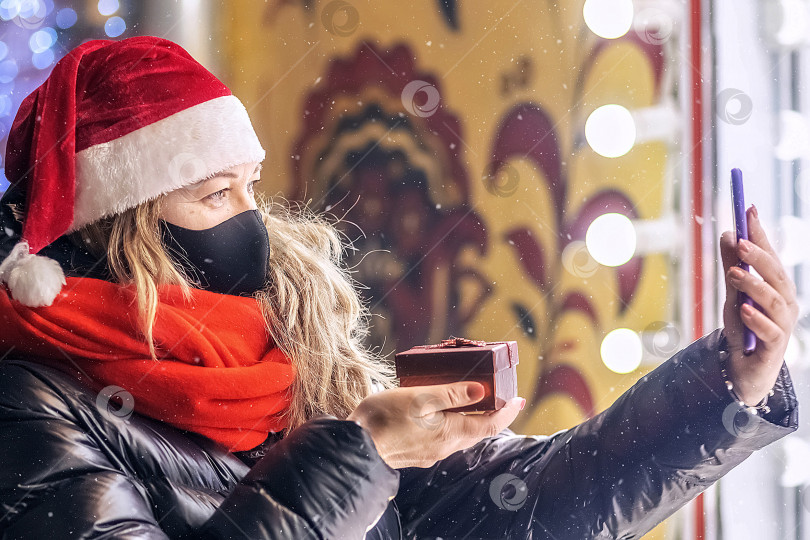 Скачать Блондинка средних лет в шляпе Санта-Клауса и защитной маске поздравляет друзей с Рождеством с помощью видео на смартфоне. Рождественские каникулы,шопинг фотосток Ozero