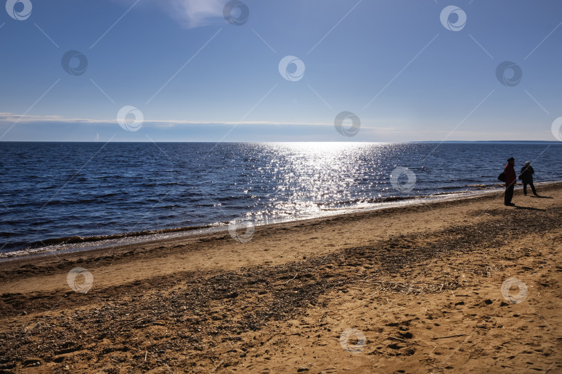 Скачать Песчаный пляж в солнечный день, синяя вода, свет Солнца отражается в воде, двое гуляющих людей фотосток Ozero