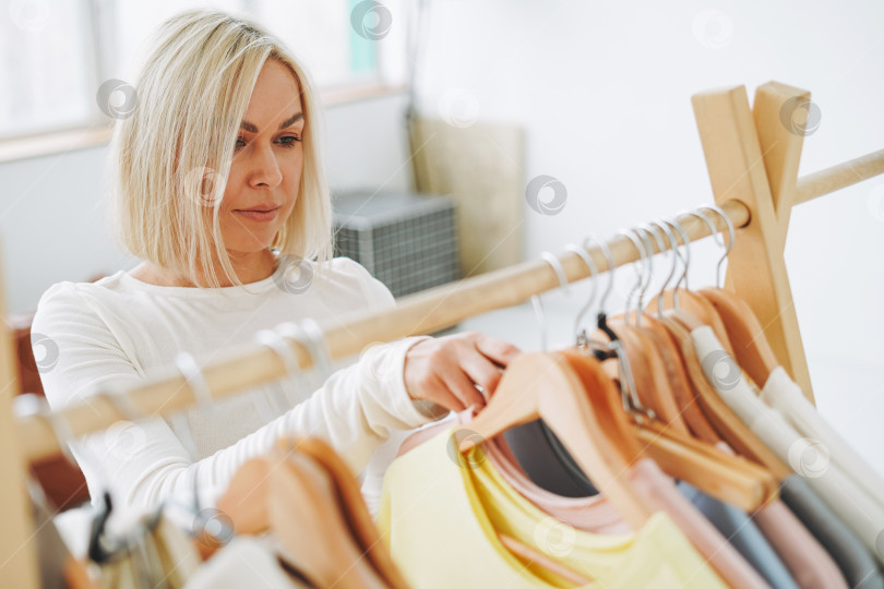 Скачать Молодая блондинка-продавец-стилист выбирает одежду на вешалке в светлой студии фотосток Ozero