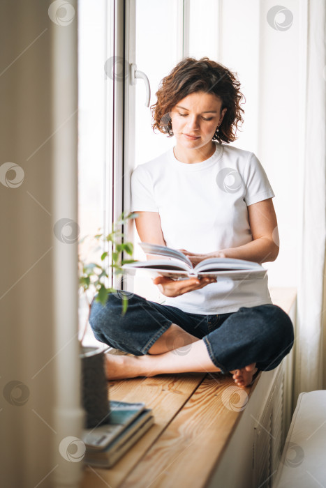 Скачать Молодая брюнетка в повседневной одежде читает книгу, сидя на подоконнике дома фотосток Ozero