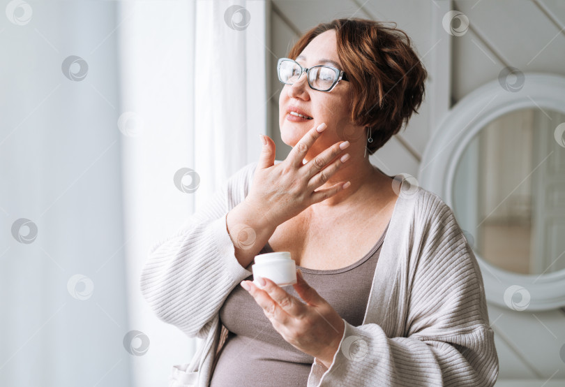 Скачать Улыбающаяся женщина средних лет плюс размер с дневным кремом у окна дома фотосток Ozero