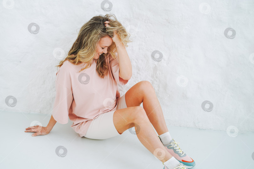 Скачать Молодая улыбающаяся блондинка в спортивной одежде сидит на фоне белой стены фотосток Ozero