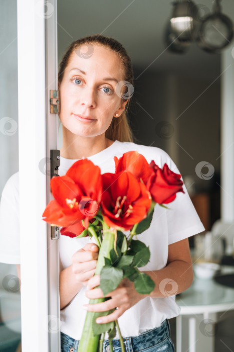 Скачать Портрет молодой женщины в белой футболке с букетом красных цветов в руках у окна на светлой кухне дома фотосток Ozero