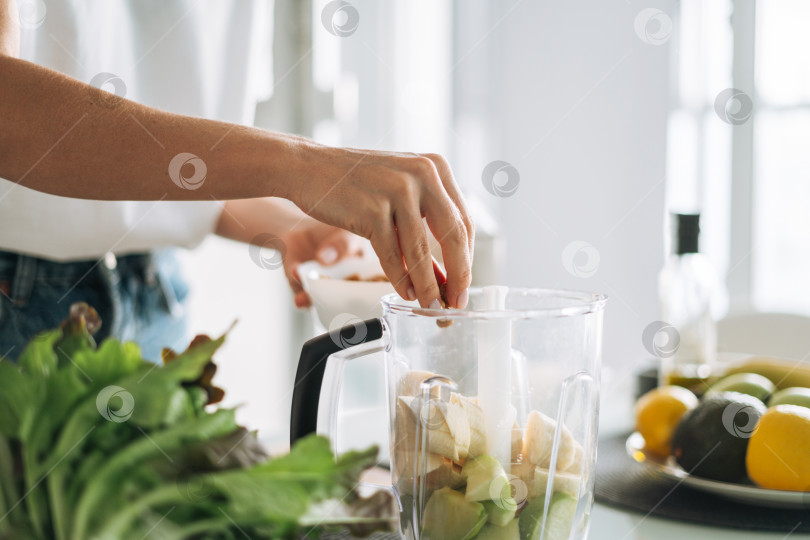 Скачать Молодая стройная женщина в белой футболке и синих джинсах готовит смузи из здоровой пищи на кухне у себя дома фотосток Ozero