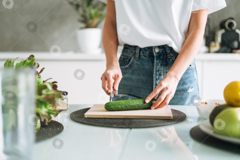 Скачать Молодая стройная женщина в белой футболке и синих джинсах готовит здоровую пищу с огурцом дома на кухне фотосток Ozero