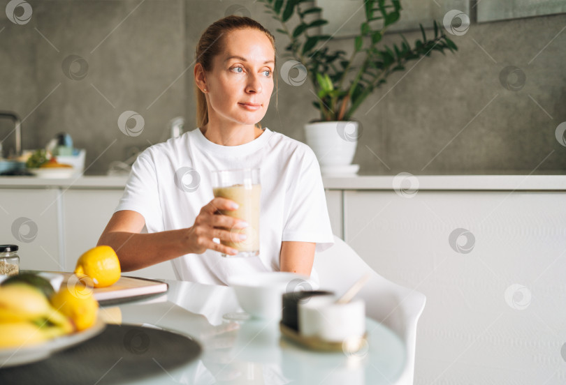 Скачать Молодая стройная женщина в белой футболке и синих джинсах пьет фруктовый коктейль "Здоровая пища" на кухне у себя дома фотосток Ozero
