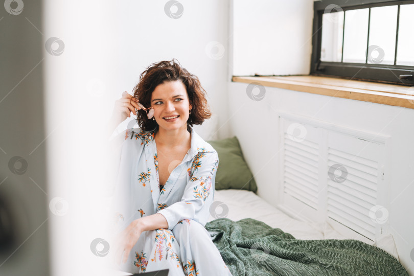Скачать Молодая брюнетка в синей пижаме делает массаж лица каменным валиком, сидя на кровати дома фотосток Ozero