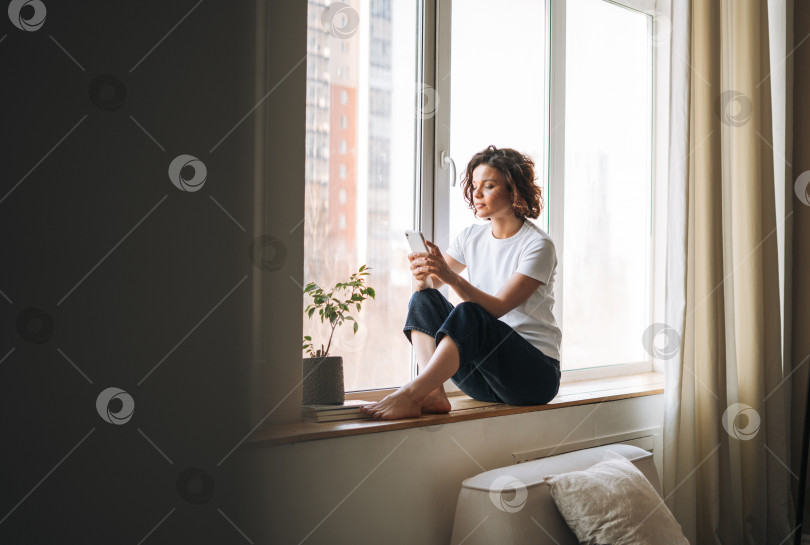 Скачать Молодая брюнетка в повседневной одежде пользуется мобильным телефоном, сидя на подоконнике дома фотосток Ozero