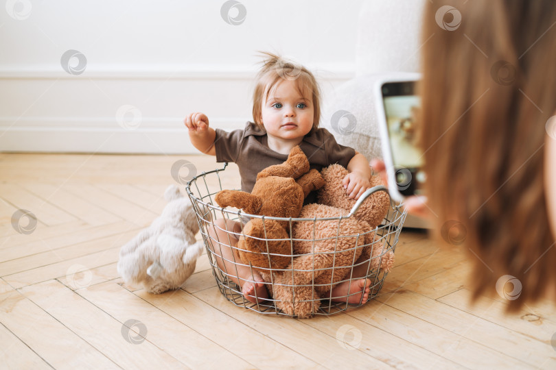 Скачать Милая малышка сидит в корзинке с мягкими игрушками, а молодая мама фотографирует ее дома на мобильный телефон фотосток Ozero
