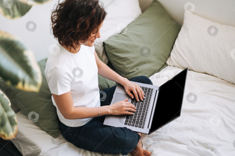 Скачать Молодая брюнетка в домашней одежде работает за ноутбуком на кровати фотосток Ozero