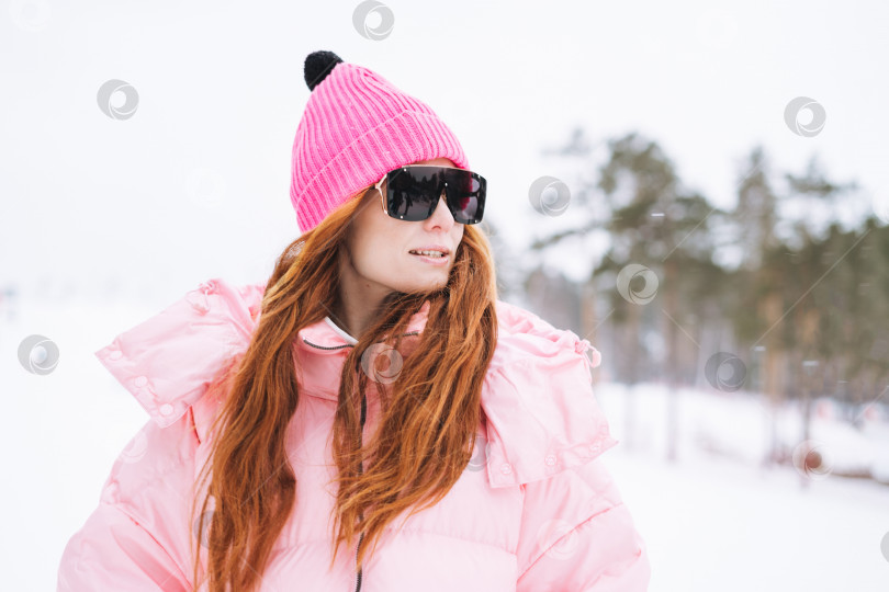 Скачать Молодая рыжеволосая женщина в розовой спортивной одежде и солнцезащитных очках на фоне зимнего снега фотосток Ozero