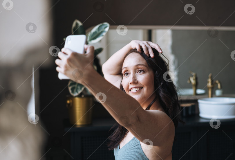 Скачать Себялюбивая женщина средних лет в нижнем белье делает селфи с помощью мобильного телефона в ванной дома фотосток Ozero