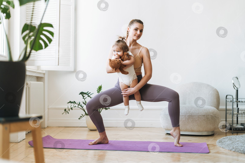 Скачать Молодая женщина, подтянутая мама с маленькой девочкой на руках, занимается фитнесом на коврике дома фотосток Ozero