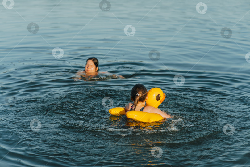 Скачать Веселая девочка развлекается в воде с желтой уткой, плавающей на поплавке, под бдительным присмотром матери фотосток Ozero