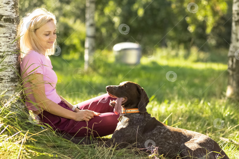 Скачать Портрет молодой светловолосой женщины, сидящей, отдыхающей на траве возле дерева в парке с охотничьей собакой породы Курц-Хаар. летние каникулы фотосток Ozero