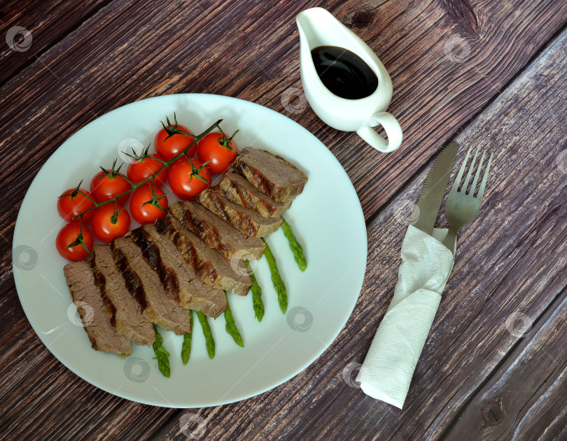 Скачать Обжаренный кусок говядины, нарезанный кусочками, отварная спаржа и помидоры черри на деревянном столе, столовые приборы в салфетке и соус рядом. фотосток Ozero